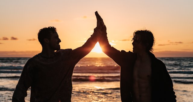 Photo of two men doing a high-five at a beach silohuetted by a sunset